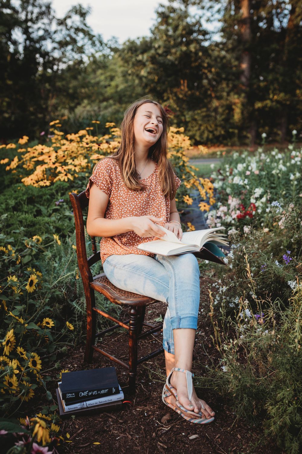 Reading outdoors in a garden filled with yellow and white flowers on a wooden chair.