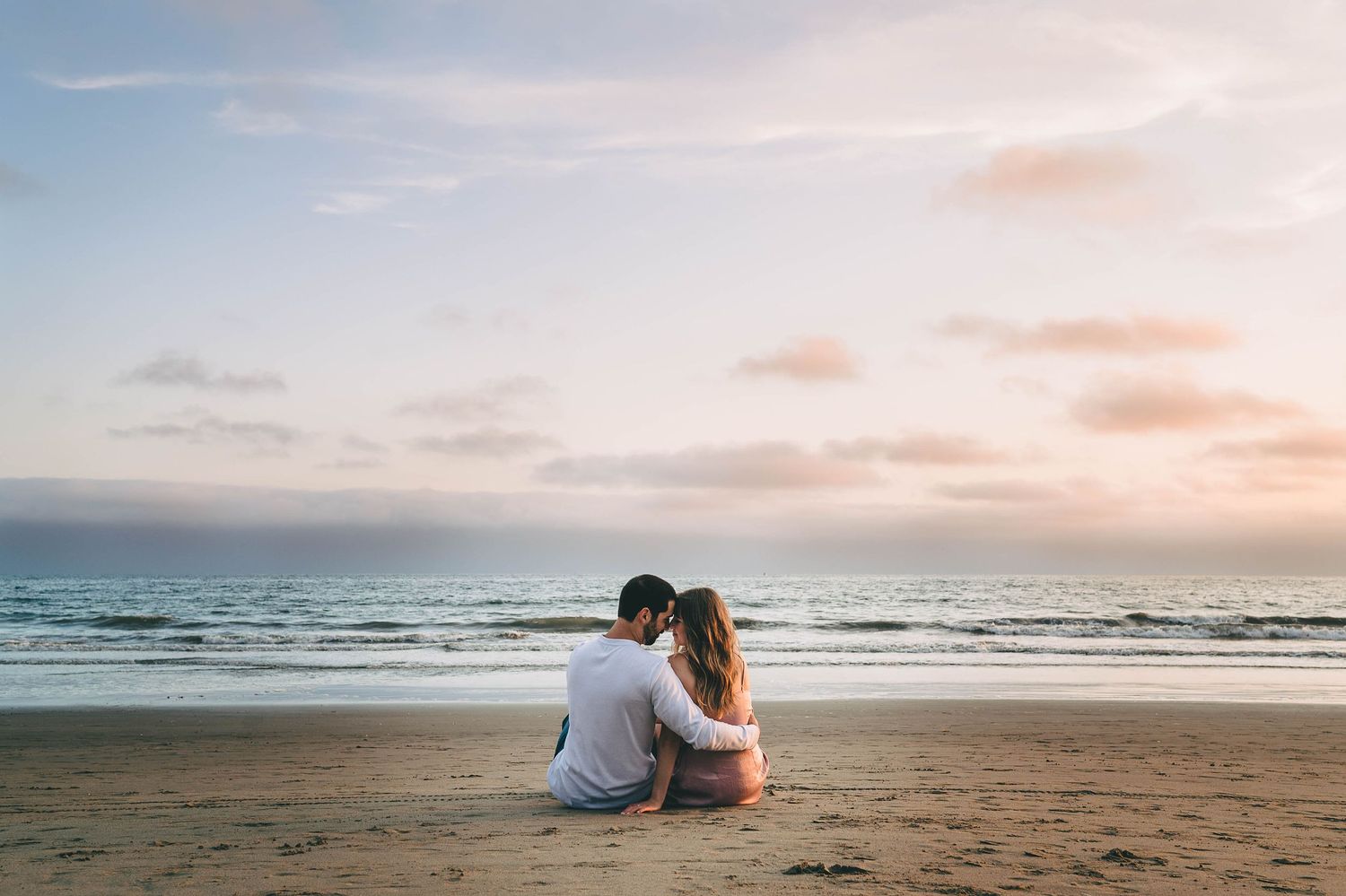 Two people sitting together on a sandy beach watching the sunset over the ocean.