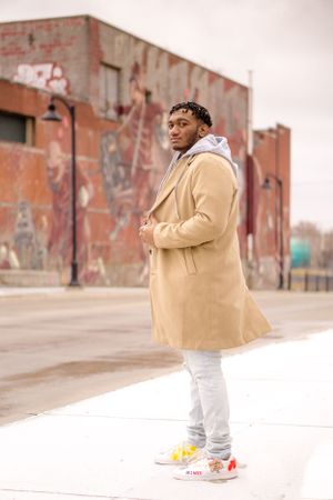 Person wearing tan coat and white pants poses on urban street with brick buildings.