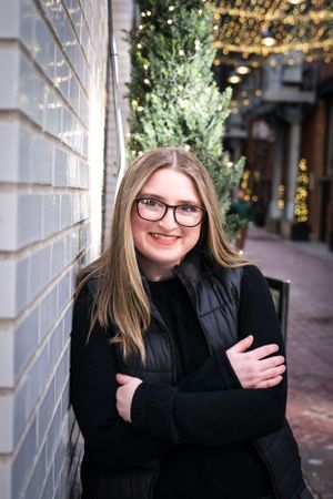 Portrait of someone in black winter coat leaning against brick wall with holiday lights.