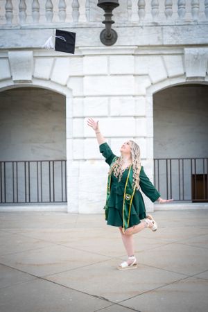 Graduate in green gown and ballet shoes tossing cap in front of white stone building.