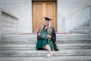 Graduate in green regalia sitting on stone steps by wooden door.