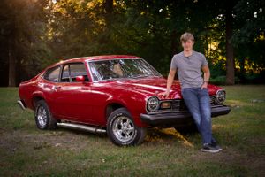 Classic red muscle car with person leaning against it in autumn setting.