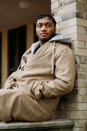 Person in beige coat sits casually on brick ledge against building exterior.