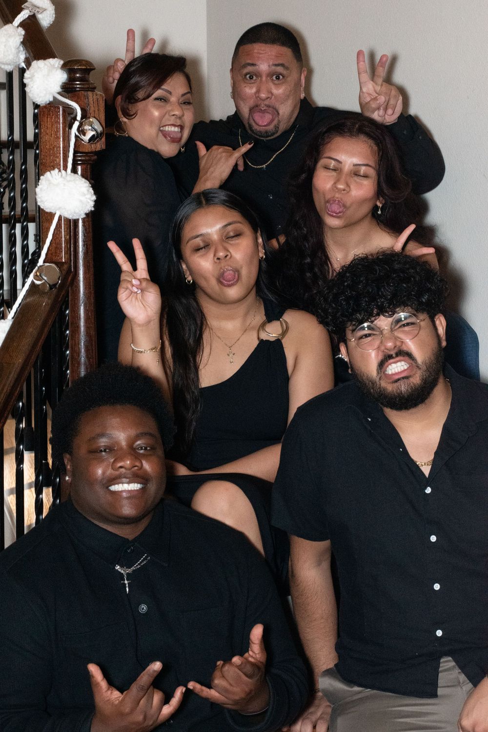 A group of friends posing together on stairs making silly faces and peace signs while wearing black outfits.