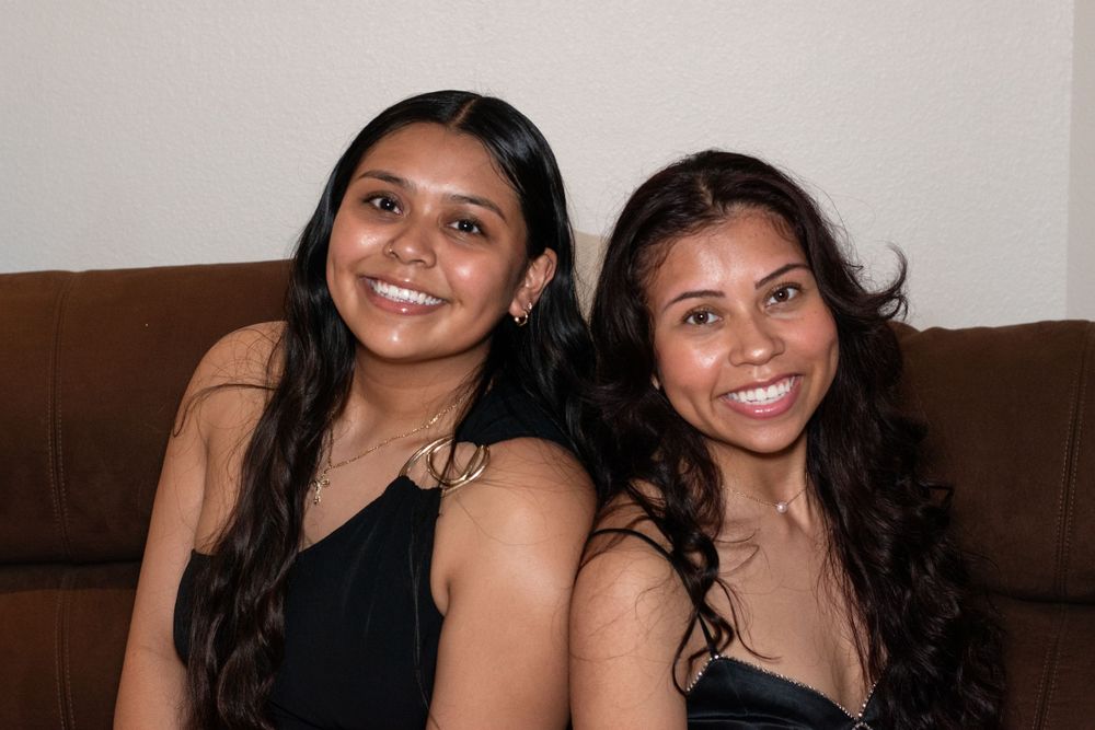 Two women in dark dresses smile while sitting on a brown couch.
