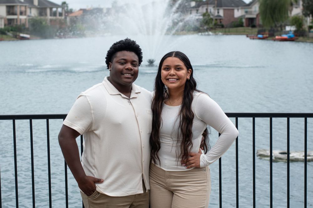 A couple poses together in neutral-colored outfits by a waterfront.