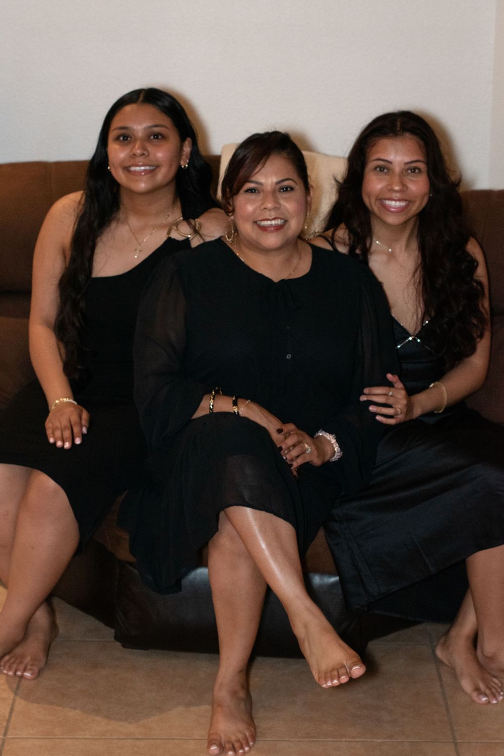 Three women in black dresses sit together on a brown couch.