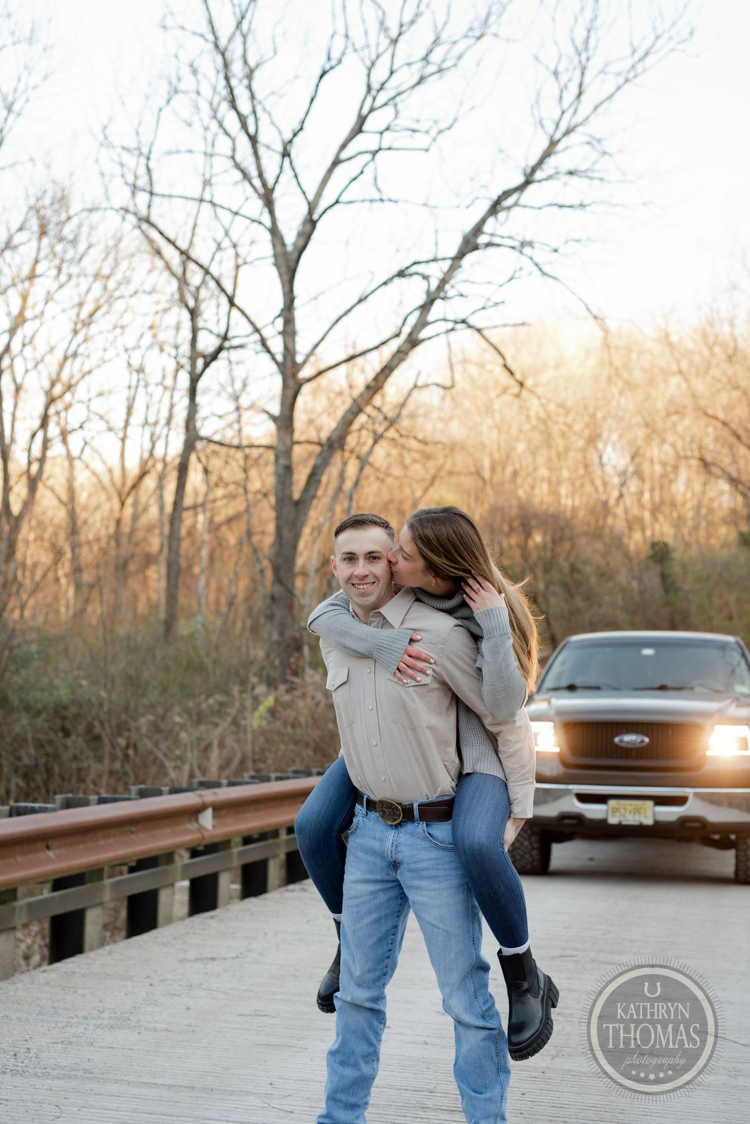 Joyful couple sharing piggyback ride on wooden bridge with truck lights glowing in background.
