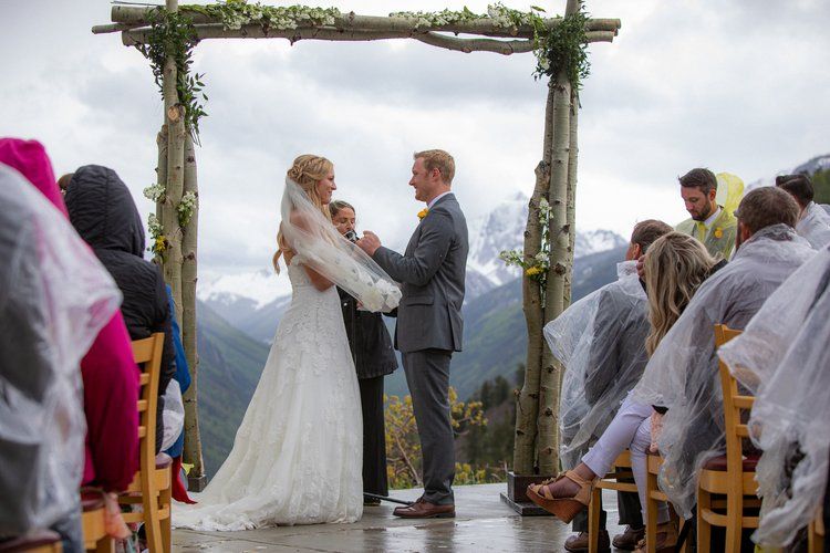 A mountain wedding ceremony under a rustic wooden arch with guests seated outdoors against a scenic backdrop.