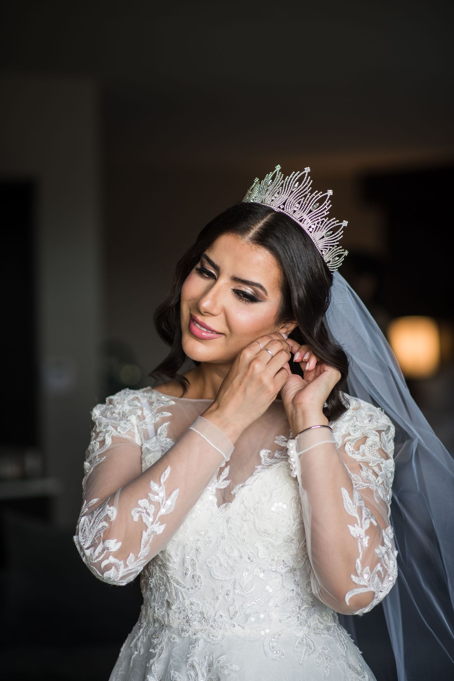Beautiful bride wearing a jeweled crown adjusts her earring in her lace wedding gown.