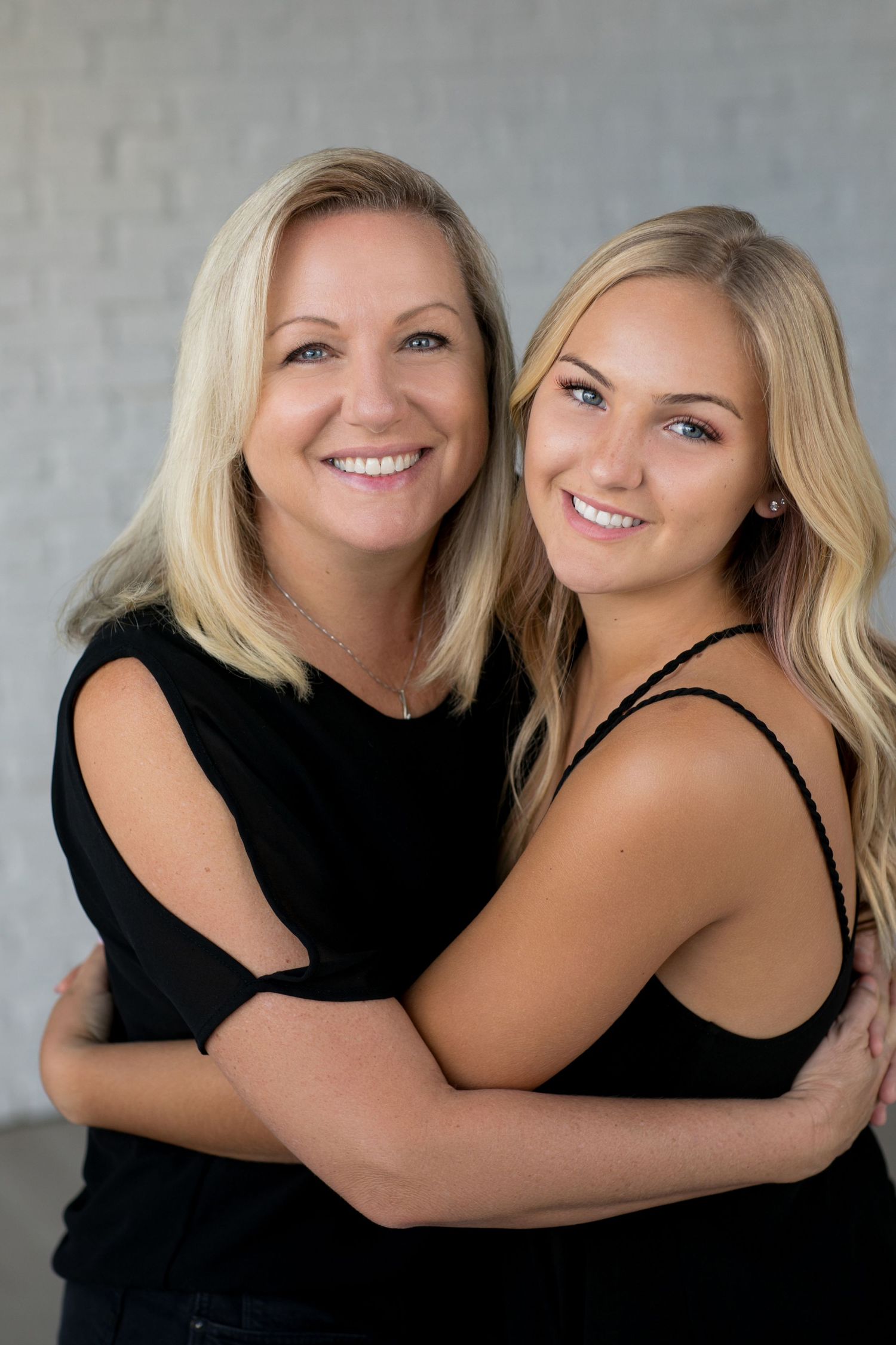 Two people in black tops embrace while posing for a close portrait against a gray background.