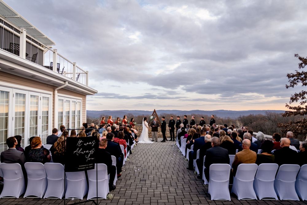 Outdoor wedding ceremony setup with white chairs and guests overlooking a scenic waterfront view under cloudy skies.