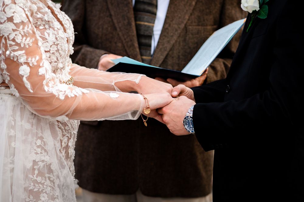 Close-up of wedding ceremony hands exchanging rings with bride wearing lace sleeve dress.