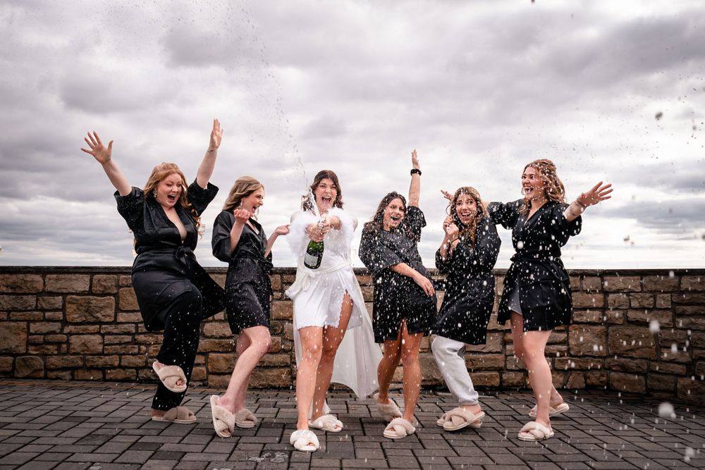 Wedding party jumping and celebrating on stone wall with cloudy sky backdrop.