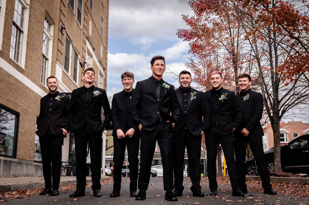 Groomsmen in dark suits line up outdoors against brick building backdrop.