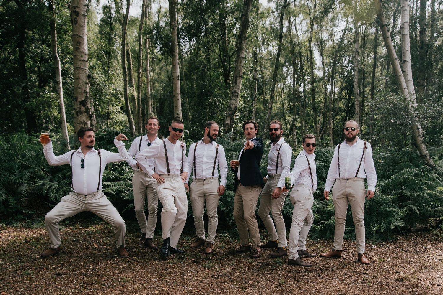Wedding party in suspenders and light-colored attire posing playfully in a forest setting.