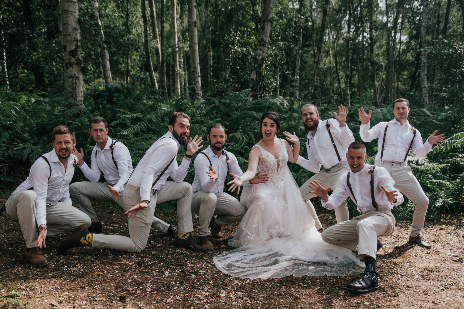 Wedding party in white and beige attire posing playfully on forest path.