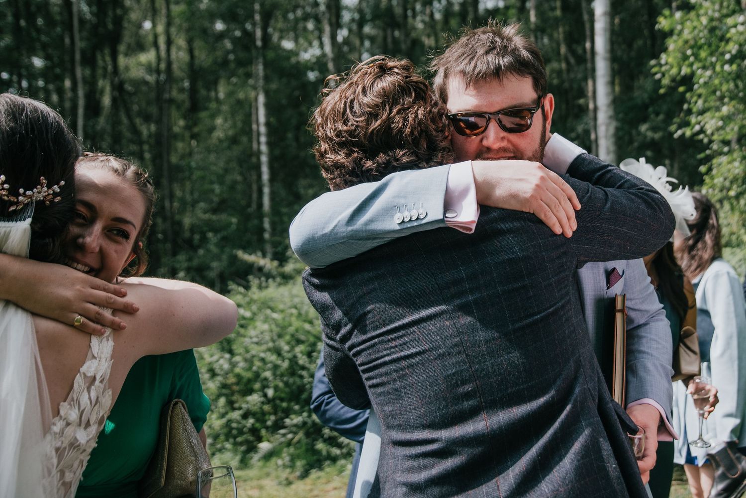 Emotional wedding guests share embraces during outdoor forest celebration.