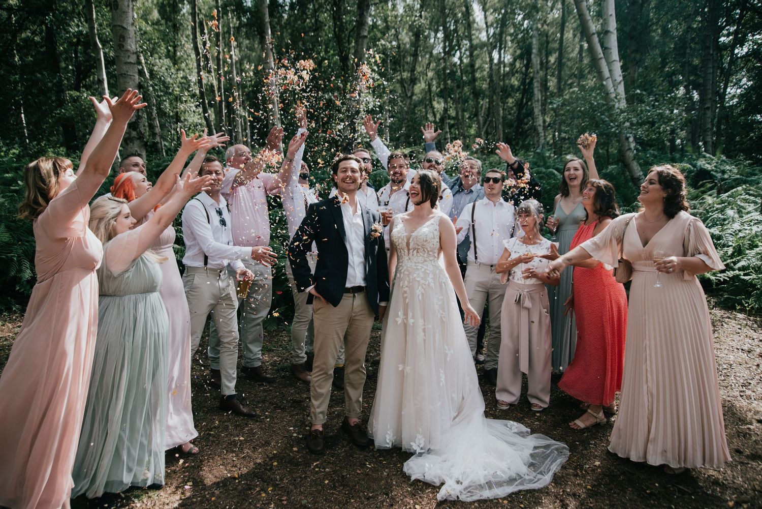 Wedding celebration with joyful group throwing confetti in a wooded outdoor setting.