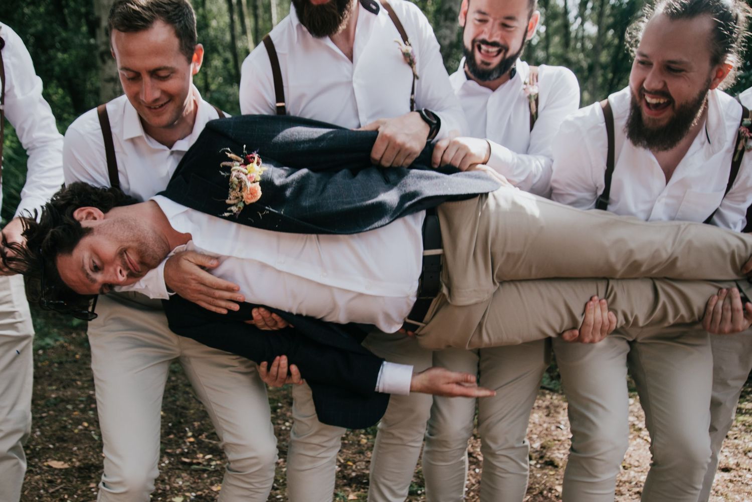 Groomsmen in white shirts and suspenders carrying someone horizontally in woodland setting.