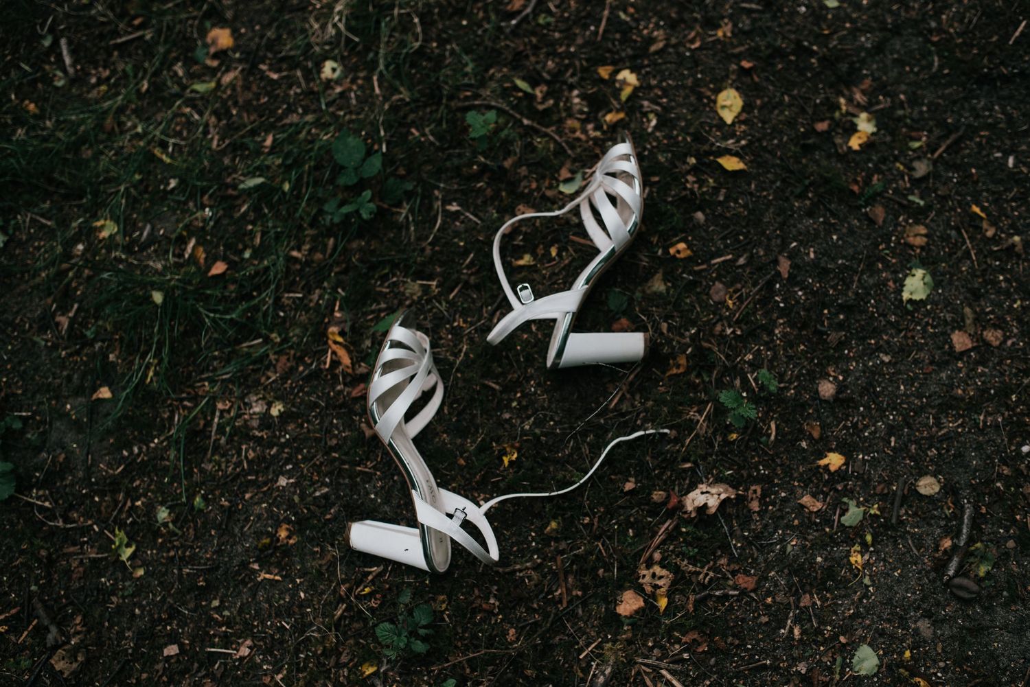 A pair of white strappy wedding heels placed on dark ground outdoors.