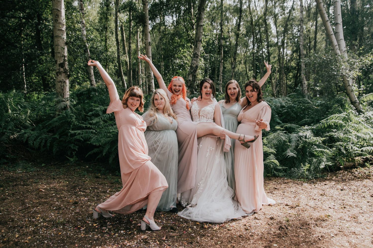 Wedding party in pastel dresses striking fun poses in a forest setting.