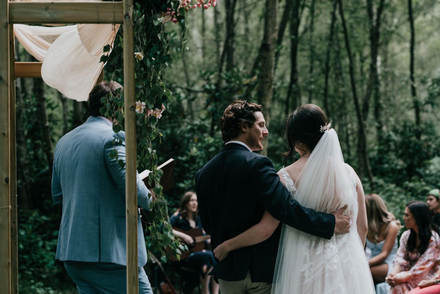 Intimate forest wedding ceremony under wooden arch decorated with flowing fabric and florals.
