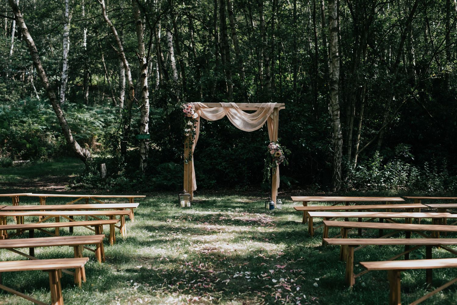 Rustic outdoor wedding ceremony setup with wooden benches and draped arch in a forest clearing.