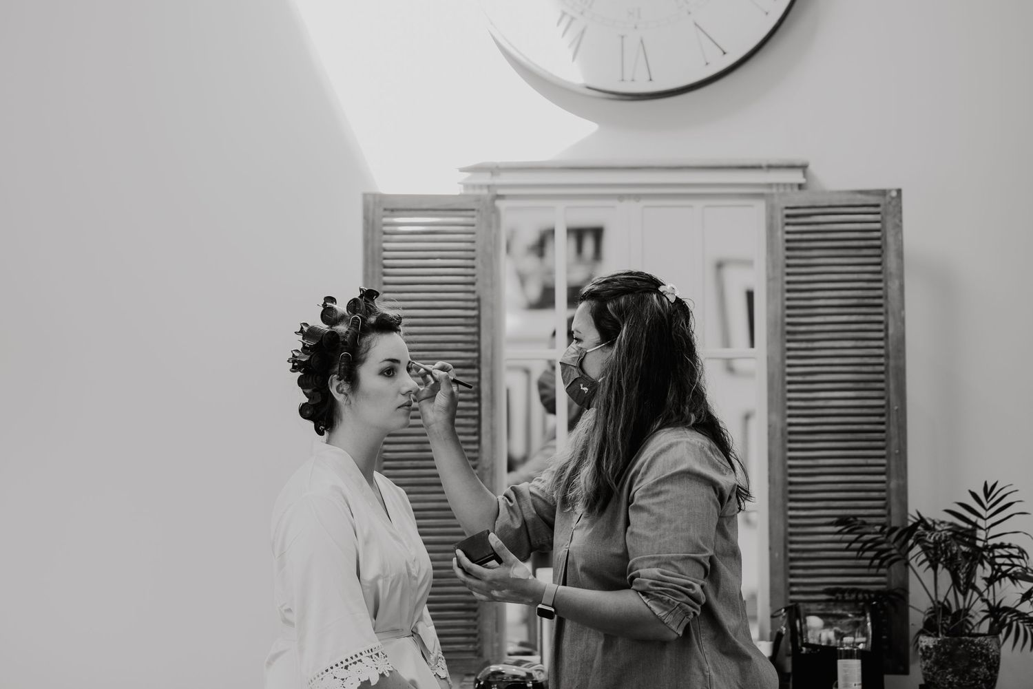 Black and white photo of wedding preparation with shuttered doors and wall clock in background.