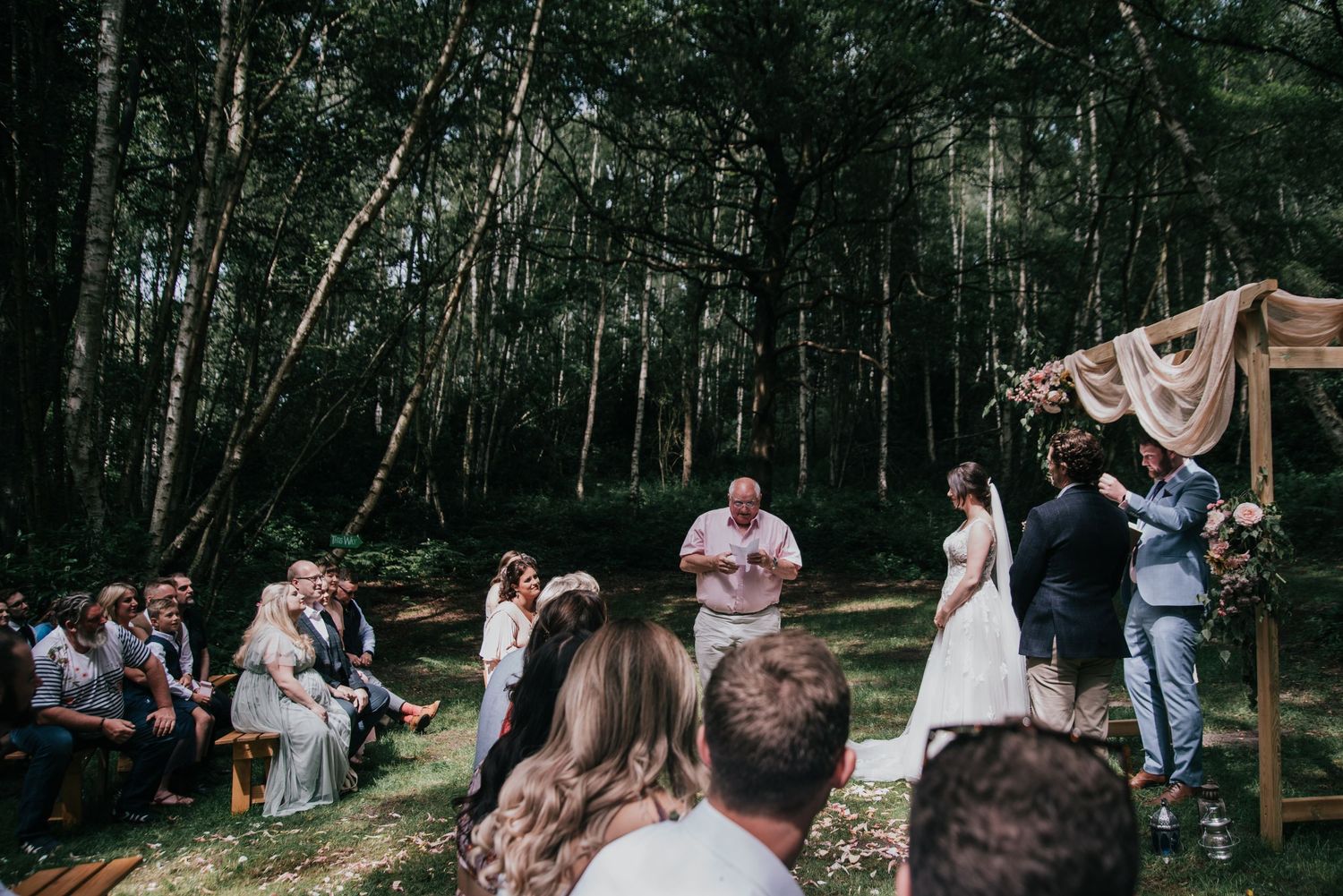Outdoor forest wedding ceremony with guests seated beneath tall trees and a draped fabric arch.