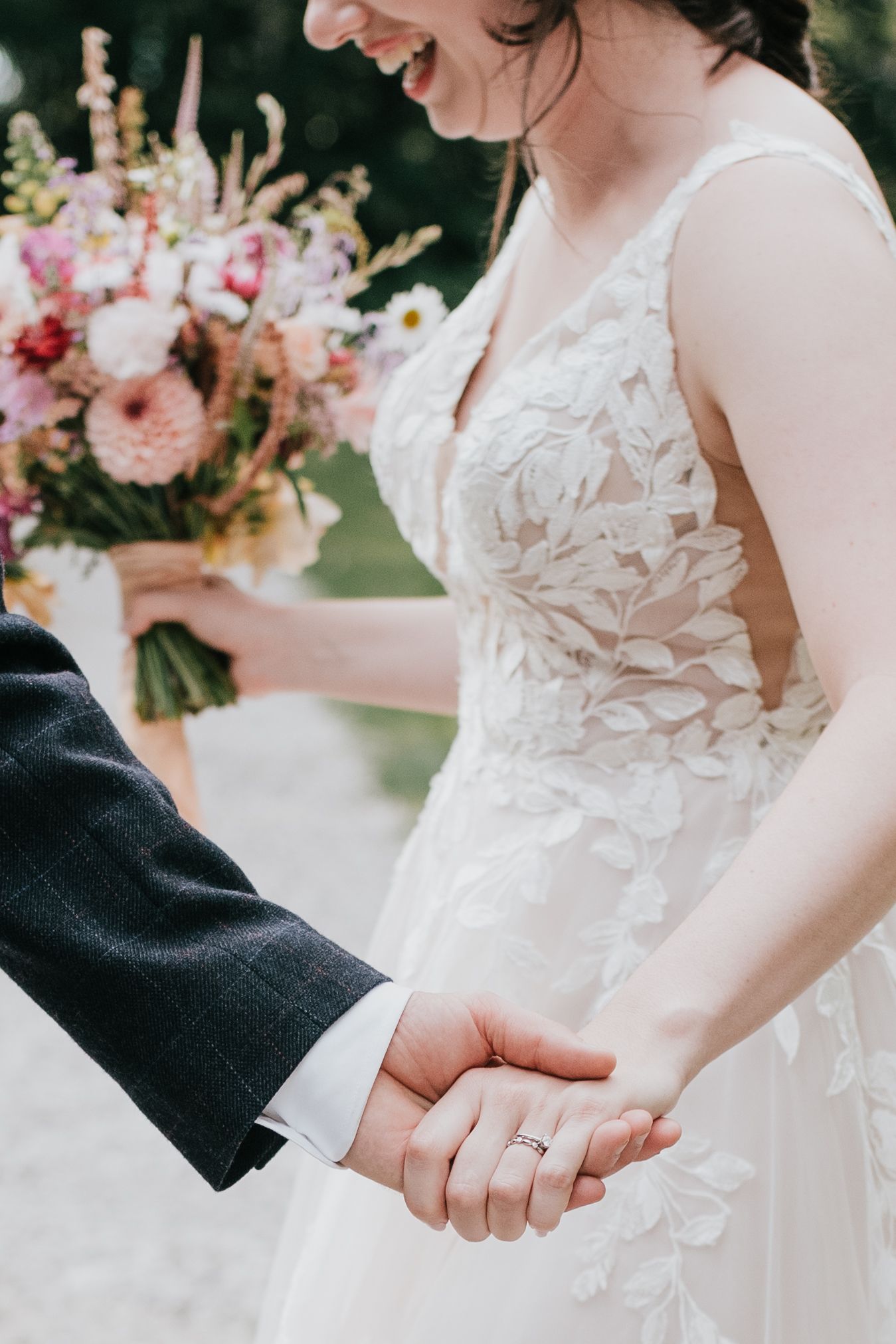 Close-up of couple holding hands with detailed lace wedding dress and pink floral bouquet visible.