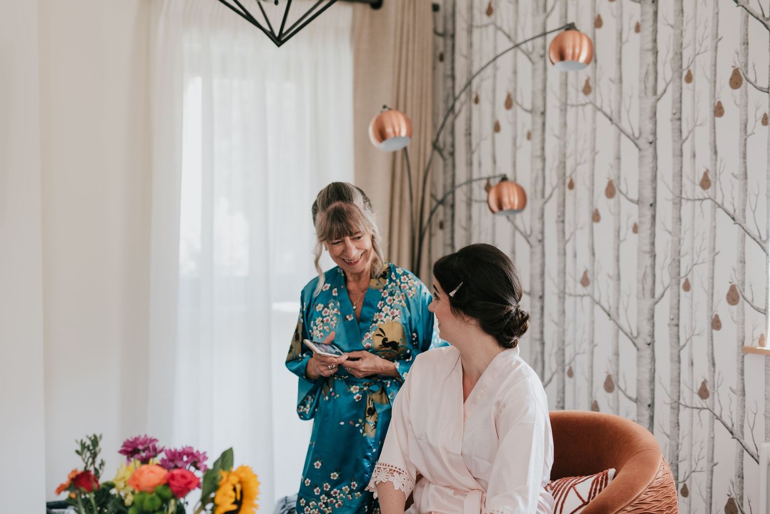 Two people in morning wedding preparation wearing robes in a room with birch wallpaper.