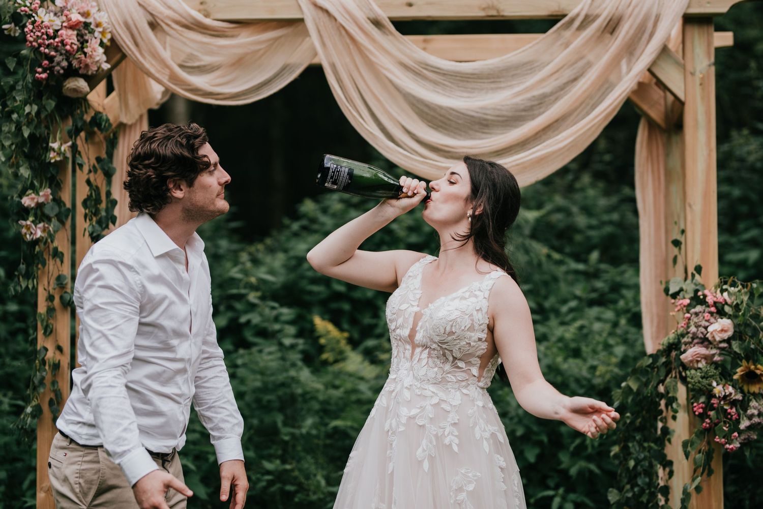A joyful moment under a draped wooden arch decorated with flowers at an outdoor wedding ceremony.