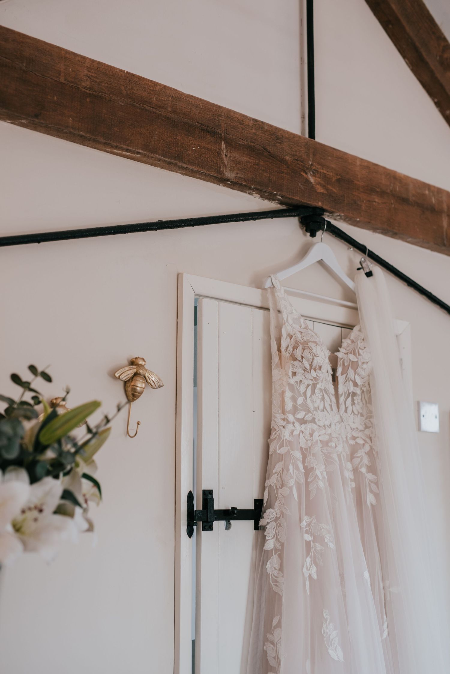 Delicate lace wedding dress hanging on display against white wall with exposed wooden beams.