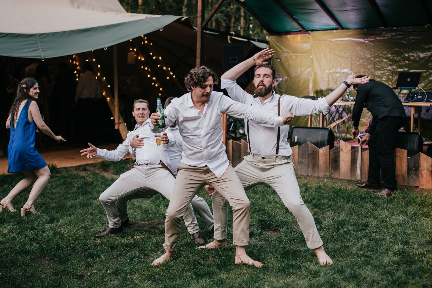 Groomsmen perform synchronized dance moves on the grass at an outdoor wedding reception.