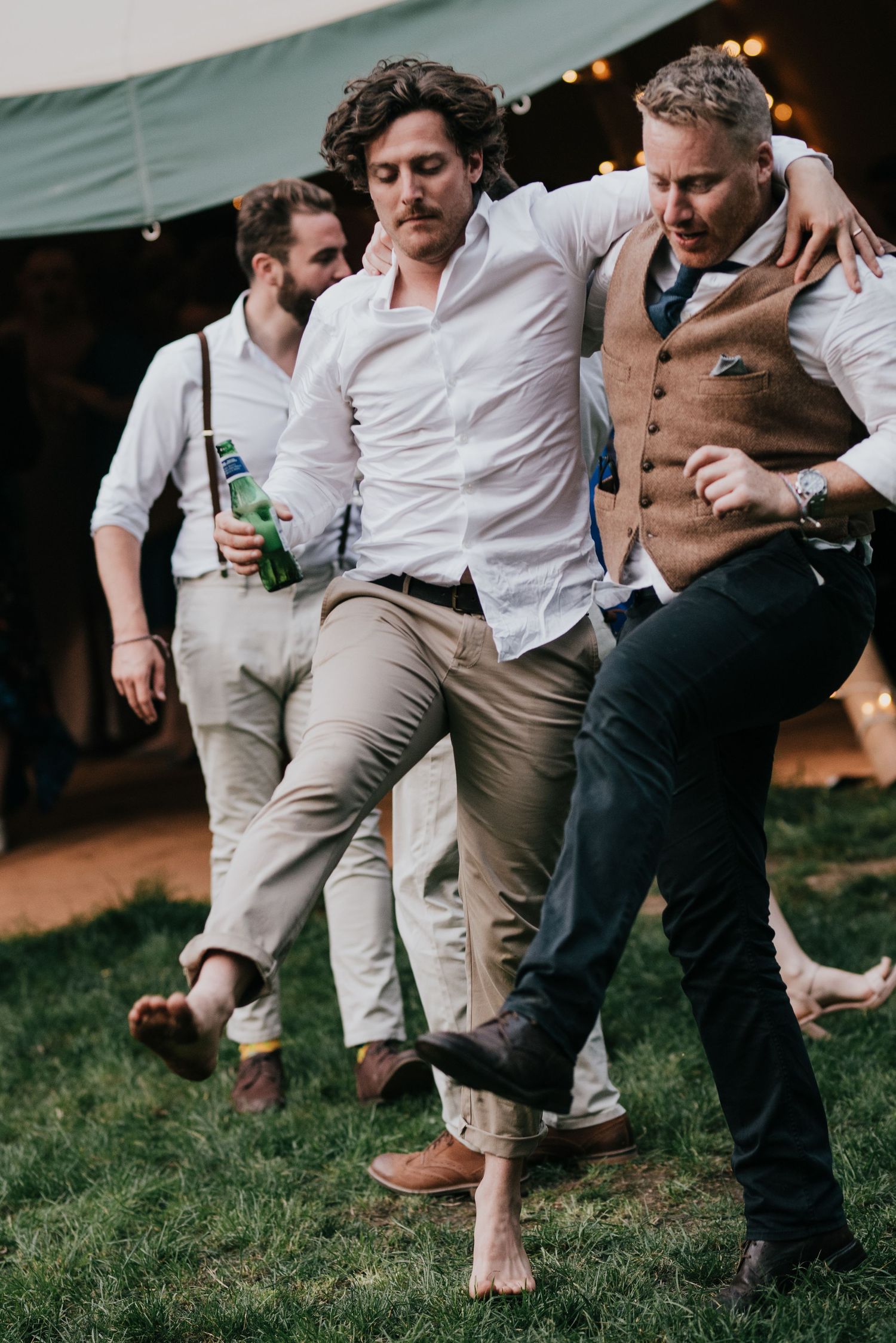 Three groomsmen in casual attire dancing and celebrating at an outdoor wedding reception.