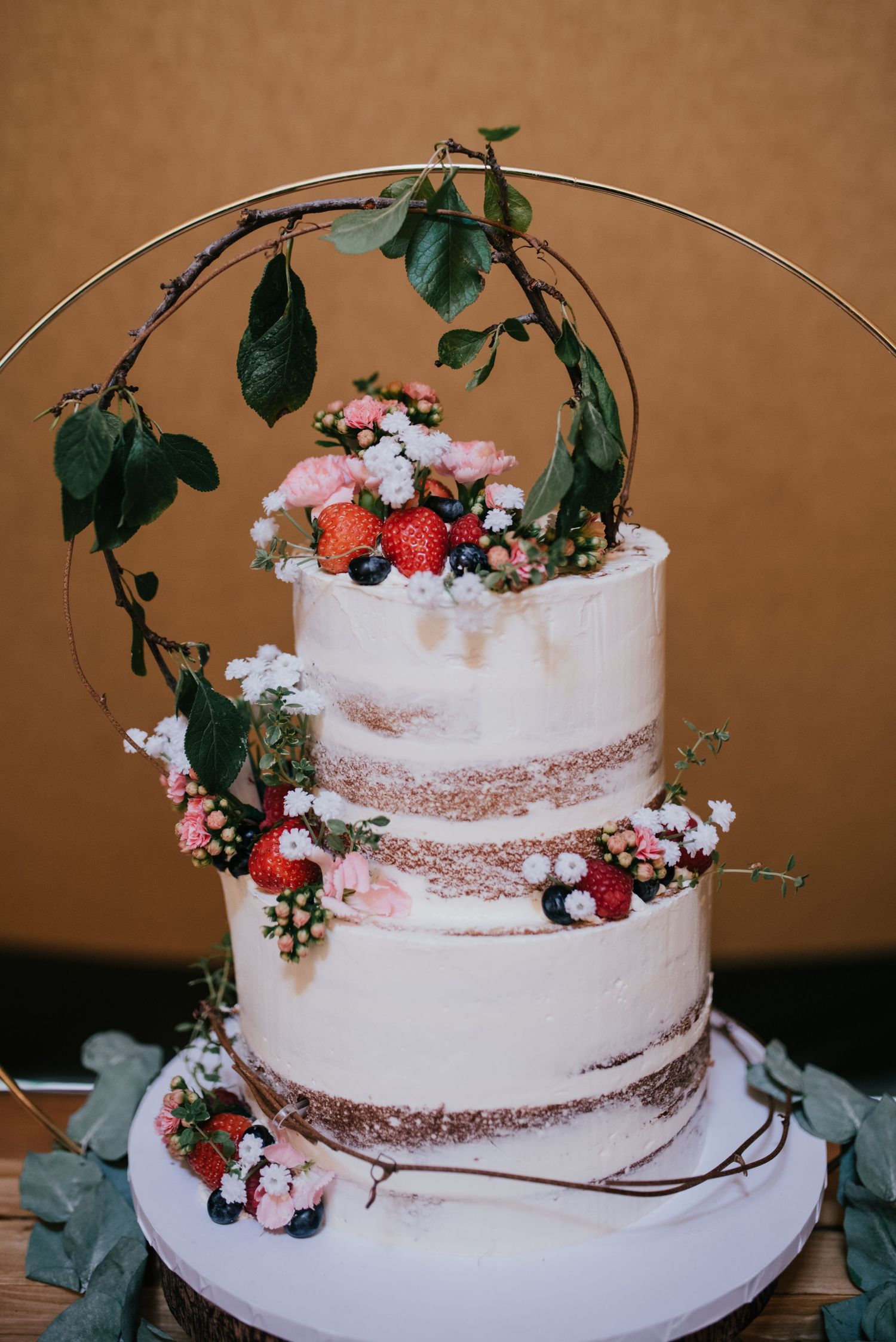 Semi-naked wedding cake decorated with fresh strawberries, flowers and green foliage arch.