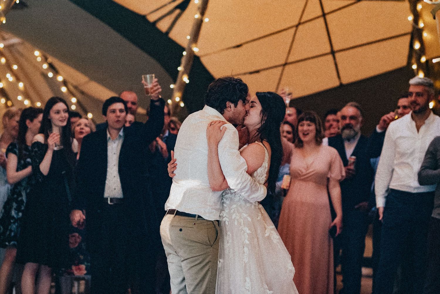 Romantic first dance under twinkling lights at an outdoor wedding reception with guests watching.