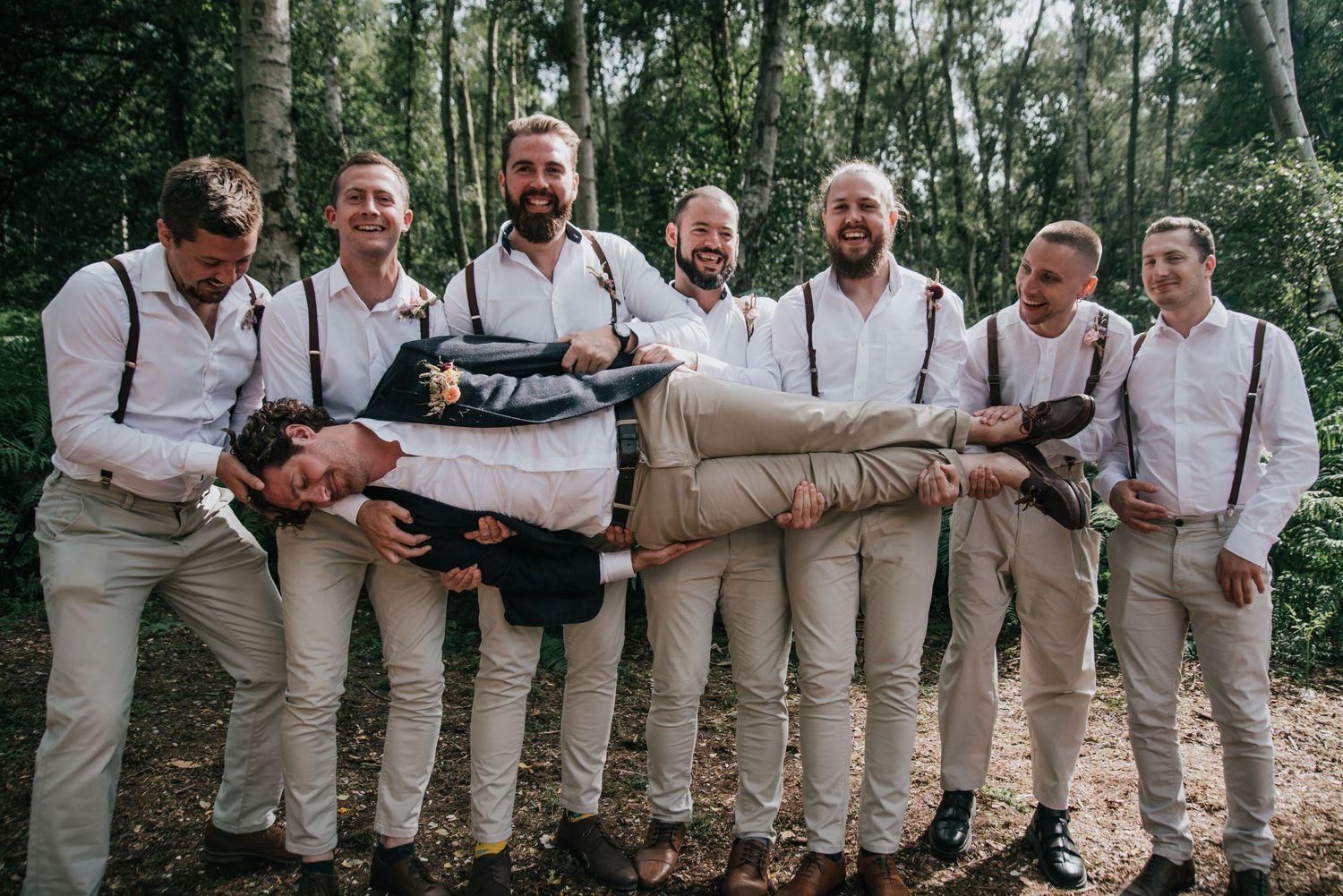 Group in matching suspenders and khakis holding someone horizontally in forest setting.