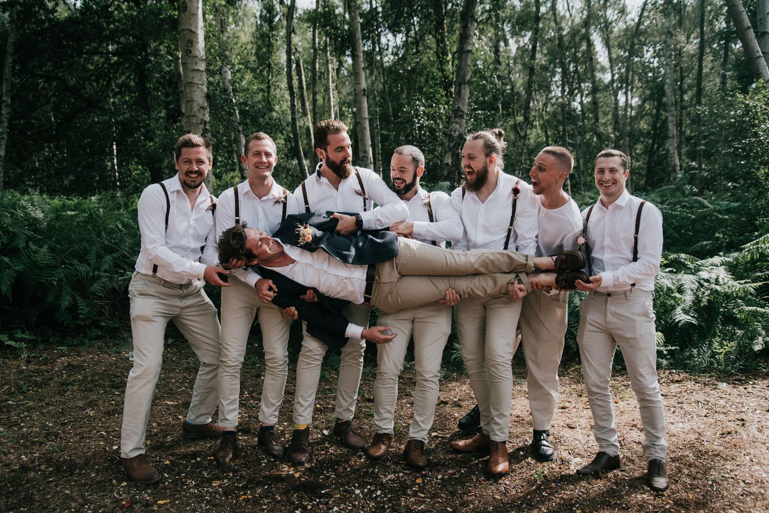 Groomsmen in coordinated outfits posing with lifted person in woodland background.