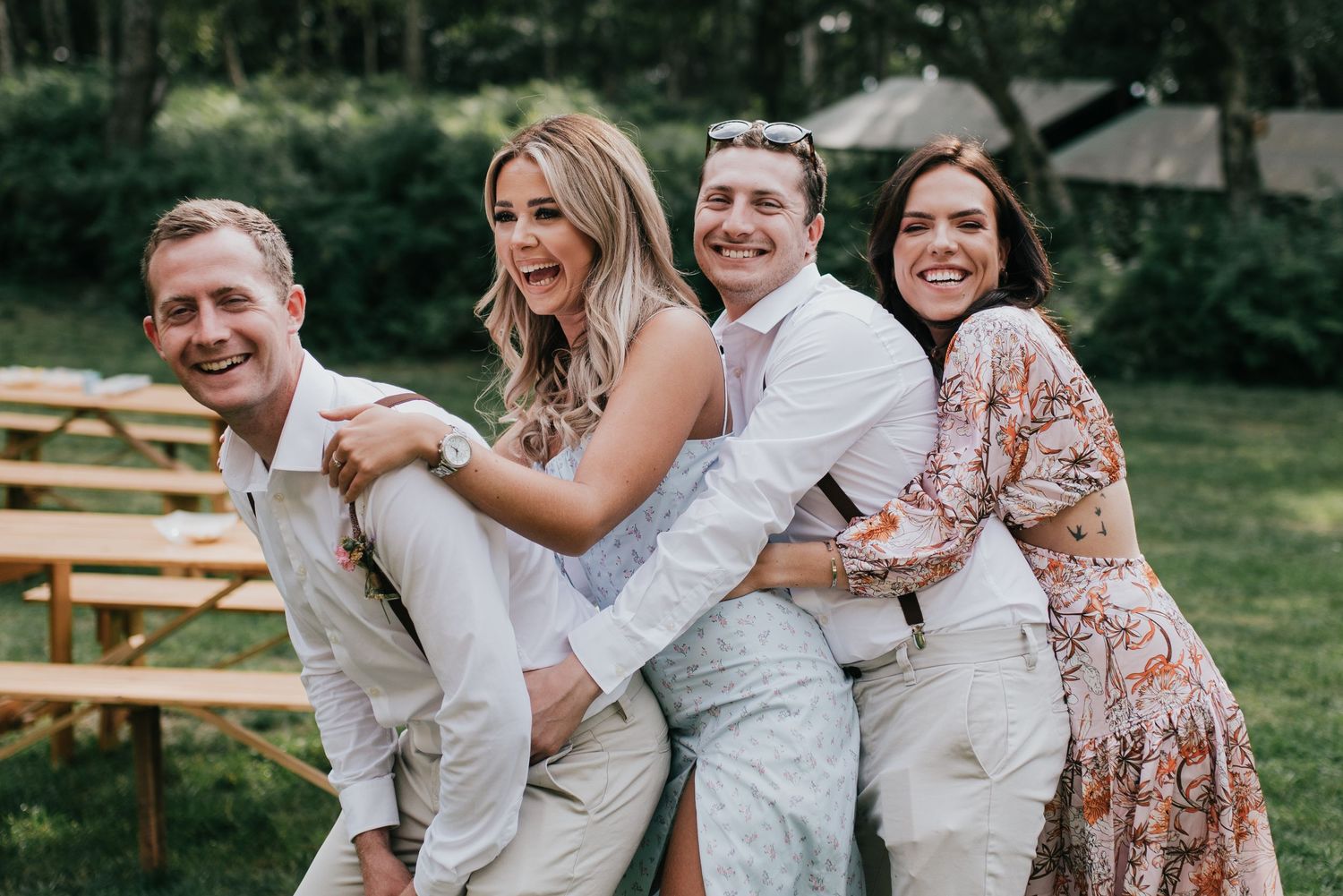 A joyful group poses together outdoors in casual attire at a wedding celebration with wooden benches in the background.