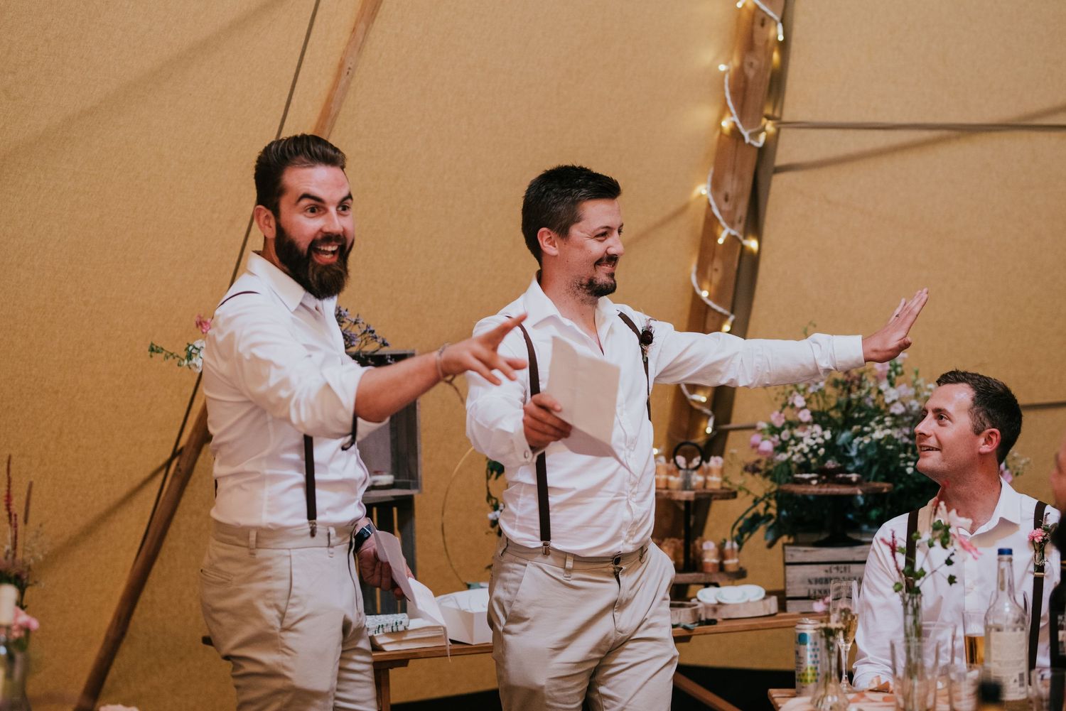Three groomsmen in light colored suspenders and dress shirts celebrate energetically inside a wedding tent.