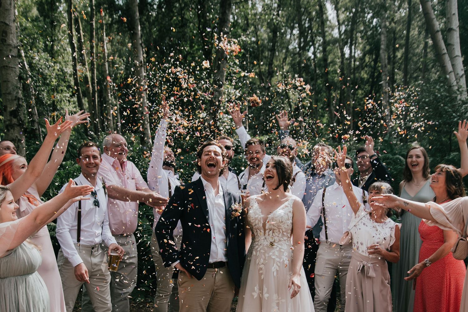 Wedding party celebrating with confetti in a sunlit forest clearing.