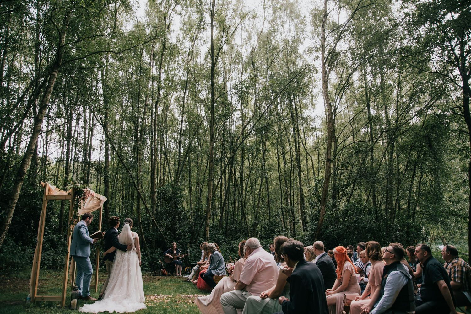 Forest wedding ceremony with wooden arch and seated guests surrounded by tall birch trees.