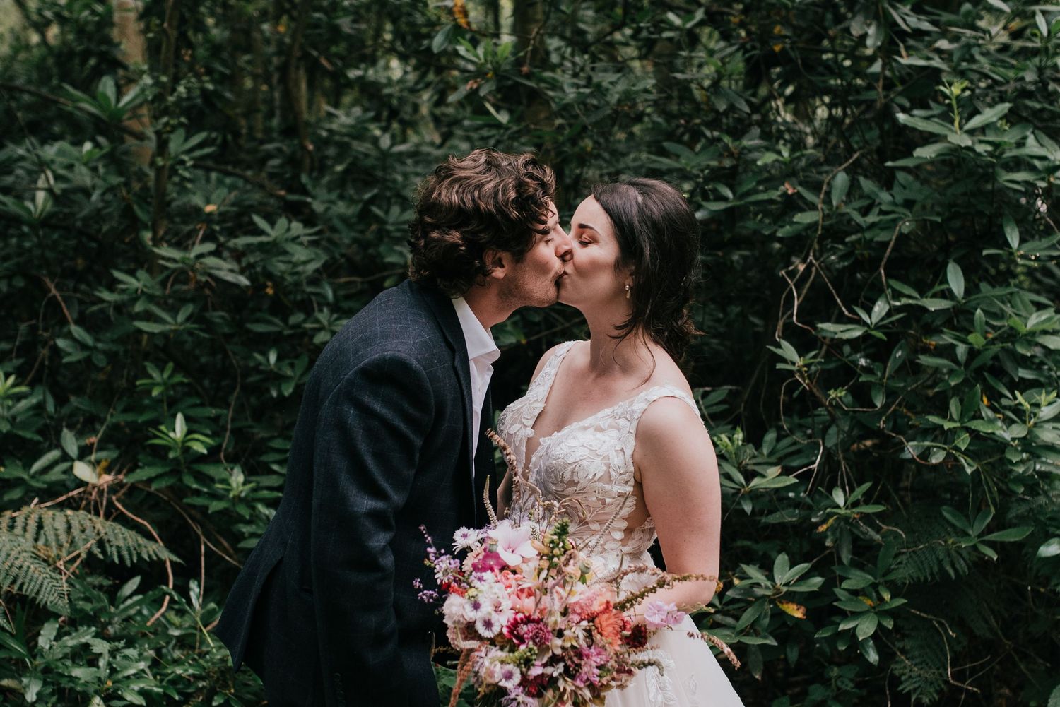 Romantic wedding couple shares a kiss in a lush garden setting while bride holds a vibrant pink bouquet.