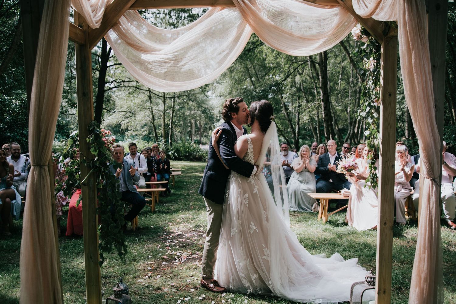 Wedding ceremony in a wooded setting with fabric-draped arch and seated guests.