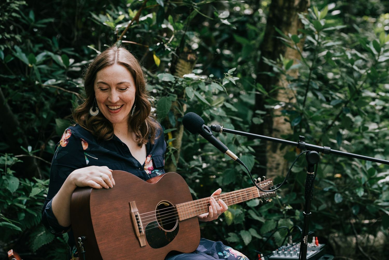 A musician playing acoustic guitar outdoors surrounded by greenery.
