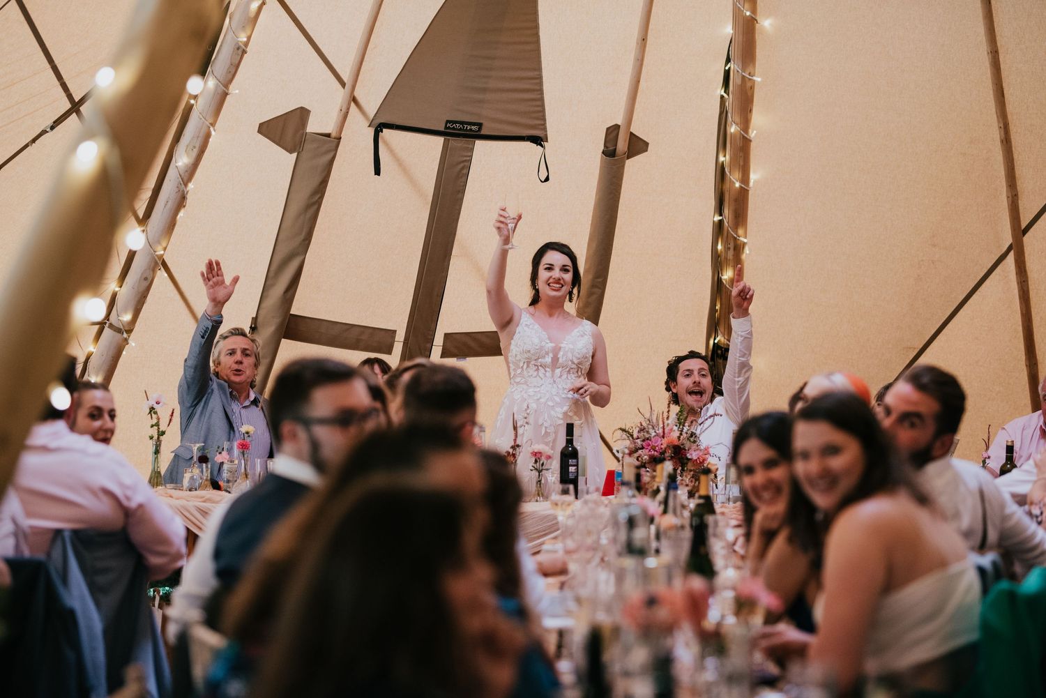 Wedding guests raise their glasses in celebration inside a warmly lit reception tent.