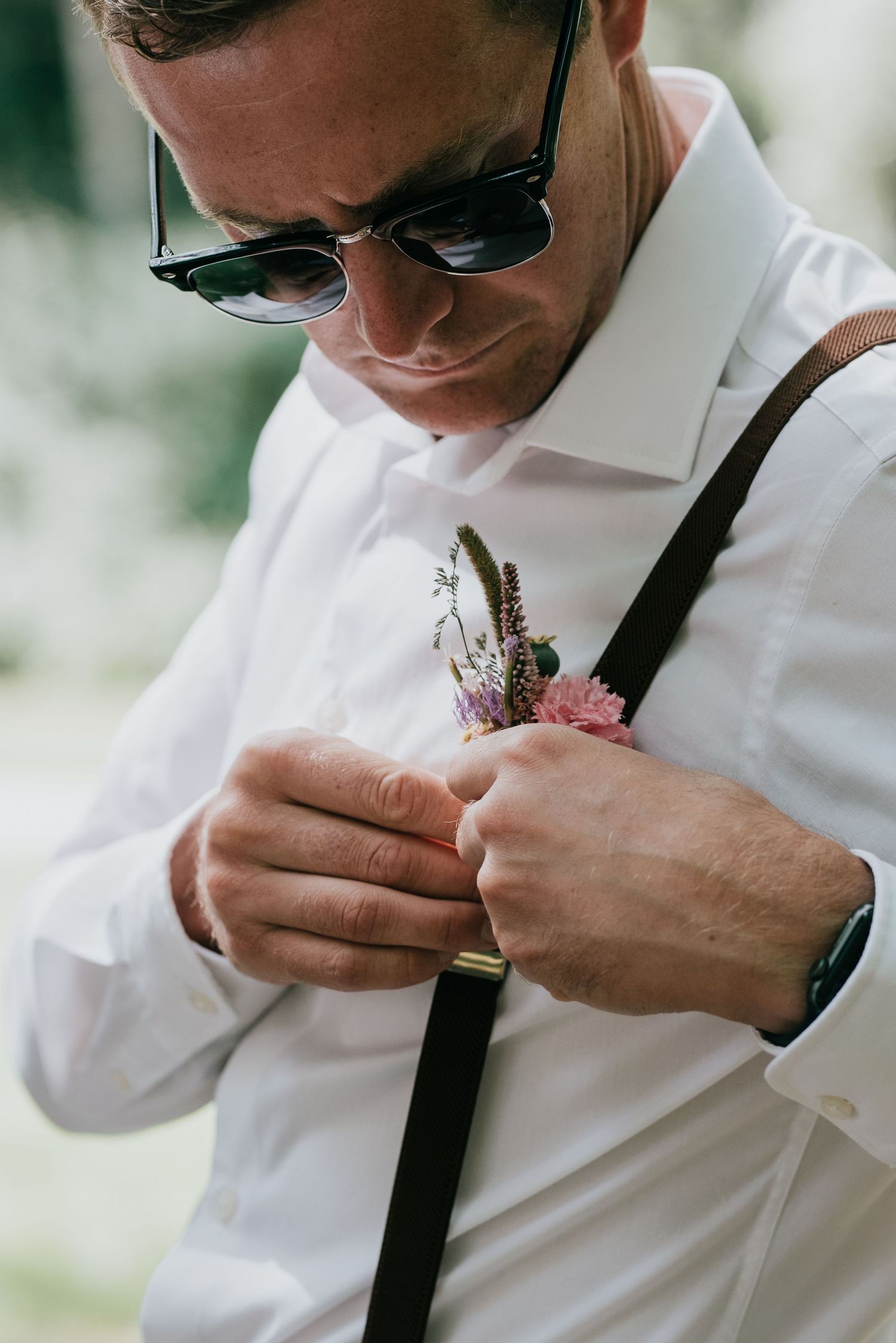 Close-up of a person in sunglasses adjusting a pink boutonniere on their white dress shirt.